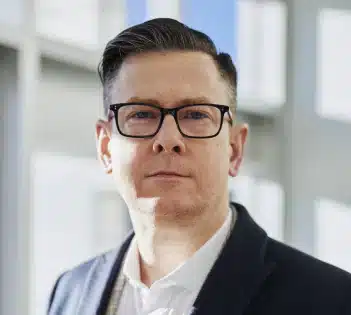 Serious-looking man wearing glasses and a dark blazer, standing in a bright office with large windows.